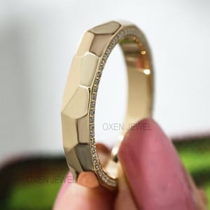 May include: A gold ring with a geometric design. The ring has a series of angled facets and a band of small, sparkling stones. The ring is held up by a person with pink nail polish.