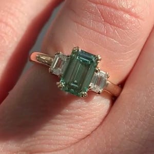May include: A gold ring featuring a large emerald-cut green gemstone flanked by two smaller baguette-cut diamonds.