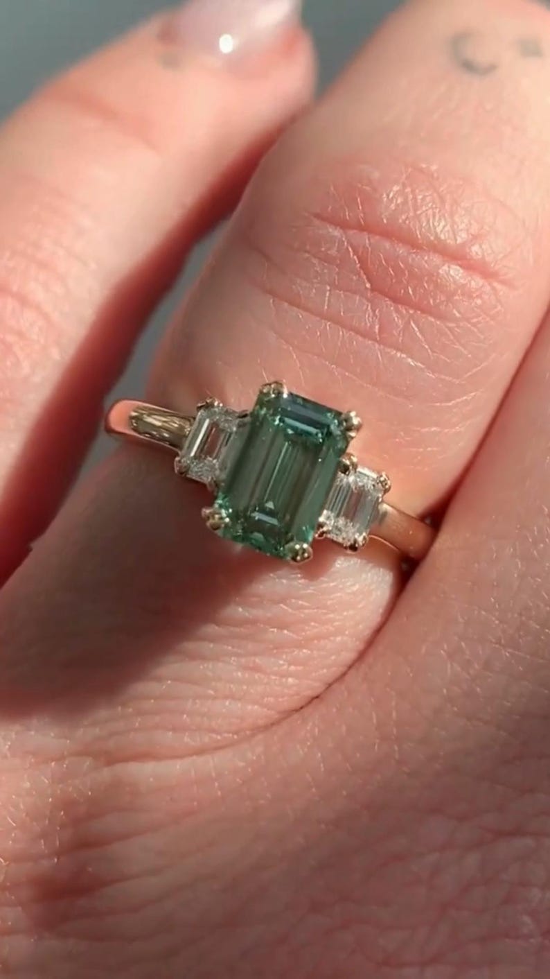 May include: A gold ring featuring a large emerald-cut green gemstone flanked by two smaller baguette-cut diamonds.