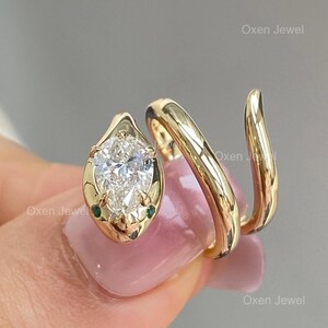 Puede incluir: Anillo de serpiente dorado con un diamante en forma de pera y dos peque&ntilde;as piedras verdes. El anillo se enrolla alrededor del dedo en espiral. El texto "Oxen Jewel" es visible.