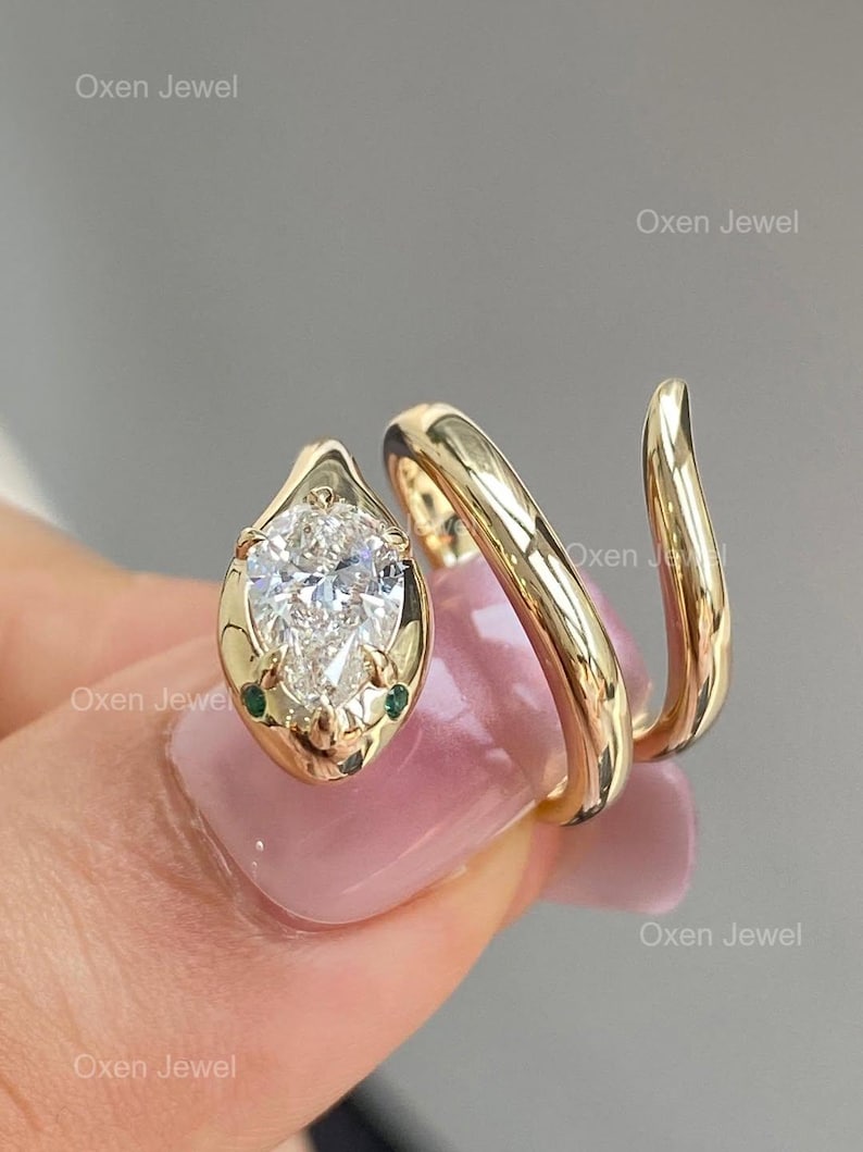 Puede incluir: Anillo de serpiente dorado con un diamante en forma de pera y dos peque&ntilde;as piedras verdes. El anillo se enrolla alrededor del dedo en espiral. El texto "Oxen Jewel" es visible.