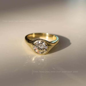 May include: A gold ring featuring a large, round, clear gemstone set in a bezel. The ring has a smooth, polished band and a classic design. The gemstone sparkles, reflecting light. The ring is displayed on a light-coloured surface.