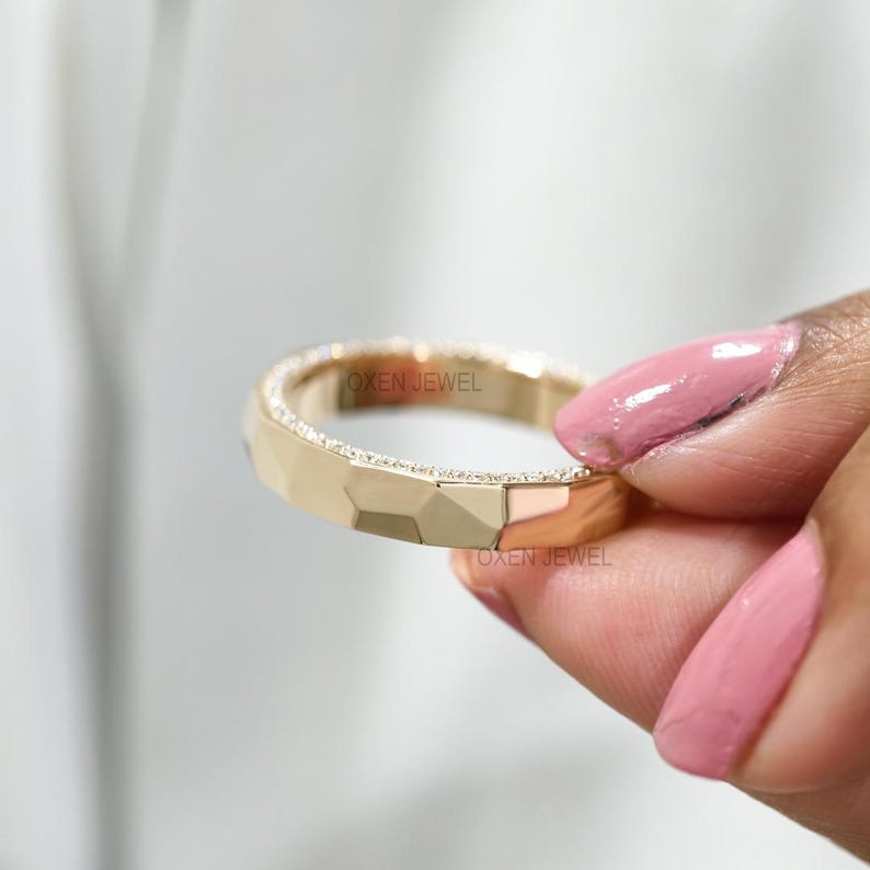 May include: A gold-colored ring with a geometric design and small, sparkling stones along the top edge. The ring is held up by a hand with pink nail polish. The text "OXEN JEWEL" is visible on the ring.
