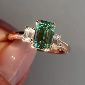 May include: A gold ring featuring a green emerald-cut gemstone and two smaller diamond accents.