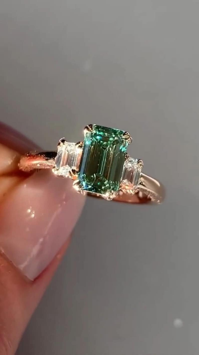 May include: A gold ring featuring a green emerald-cut gemstone and two smaller diamond accents.