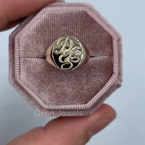 Pode incluir: Um anel de sinete dourado com uma face oval preta. A face apresenta as iniciais entrelaçadas "DG" em escrita cursiva, com um pequeno detalhe de diamante. O anel é apresentado numa caixa de veludo rosa. A marca "Oxen Jewels" é visível.