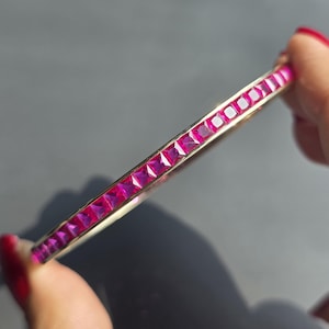 May include: A gold bracelet featuring a line of square-cut, bright pink gemstones. The bracelet is displayed against a grey backdrop, highlighting the gemstones' colour and the gold's gleam. A piece of jewellery.
