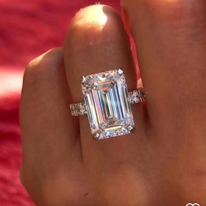 Puede incluir: Un anillo de plata con un gran diamante de talla esmeralda transparente. El diamante está engastado en un engaste de pavé con diamantes más pequeños en la banda.