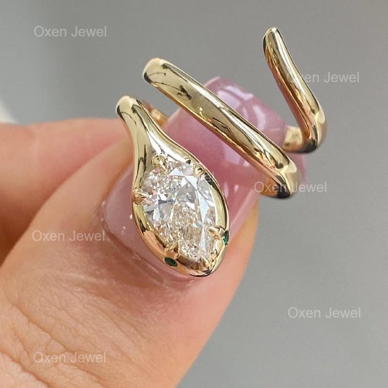 Puede incluir: Un anillo de serpiente dorado con una gema clara en forma de pera como cabeza. El anillo se enrolla alrededor del dedo, con peque&ntilde;os detalles verdes en la cabeza de la serpiente. Una joya.