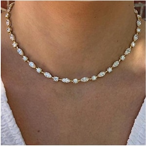 Puede incluir: Un collar dorado con piedras preciosas transparentes de talla redonda y marquesa. El collar se lleva alrededor del cuello y descansa sobre una prenda blanca. Las piedras están dispuestas en un patrón alterno.