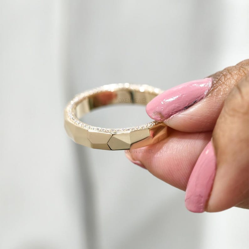 May include: A gold ring with a geometric design and a row of small, sparkling stones. The ring has a faceted surface and is held by a person with pink nail polish. The text "OXEN JEWEL" is visible.