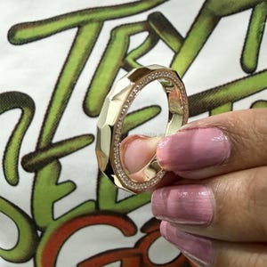 May include: A gold-colored ring with a geometric design and a band of small, clear stones. The ring has a faceted surface and is held up against a white shirt with green and orange text.
