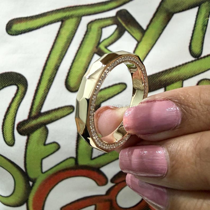 May include: A gold-colored ring with a geometric design and a band of small, clear stones. The ring has a faceted surface and is held up against a white shirt with green and orange text.