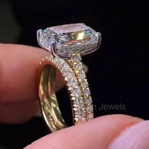 May include: A two-piece engagement ring set. The main ring features a large, rectangular-cut diamond with a halo of smaller diamonds. The band is gold and encrusted with small diamonds. The second band is a gold band with small diamonds.