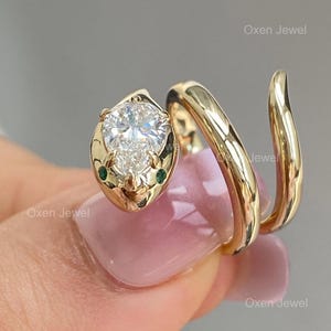 Puede incluir: Un anillo de serpiente de oro amarillo de 14k con un diamante de laboratorio con corte de pera de 1 quilate. La cabeza de la serpiente presenta el diamante y dos peque&ntilde;as piedras verdes. El cuerpo del anillo se enrolla alrededor del dedo. El texto de la imagen dice "14k yellow gold 1Ct pear cut lab diamond snake ring."