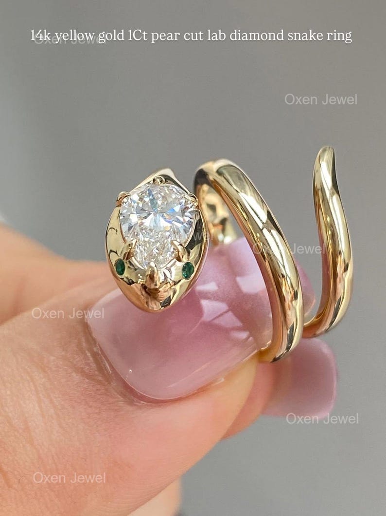 Puede incluir: Un anillo de serpiente de oro amarillo de 14k con un diamante de laboratorio con corte de pera de 1 quilate. La cabeza de la serpiente presenta el diamante y dos peque&ntilde;as piedras verdes. El cuerpo del anillo se enrolla alrededor del dedo. El texto de la imagen dice "14k yellow gold 1Ct pear cut lab diamond snake ring."