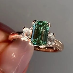 May include: A rose gold engagement ring featuring a green emerald-cut gemstone and two smaller diamond accents.