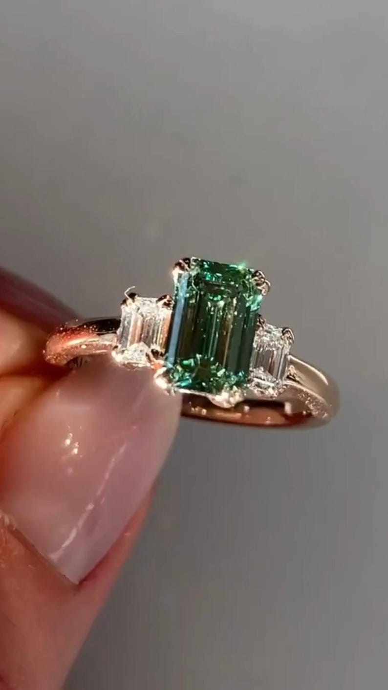 May include: A rose gold engagement ring featuring a green emerald-cut gemstone and two smaller diamond accents.