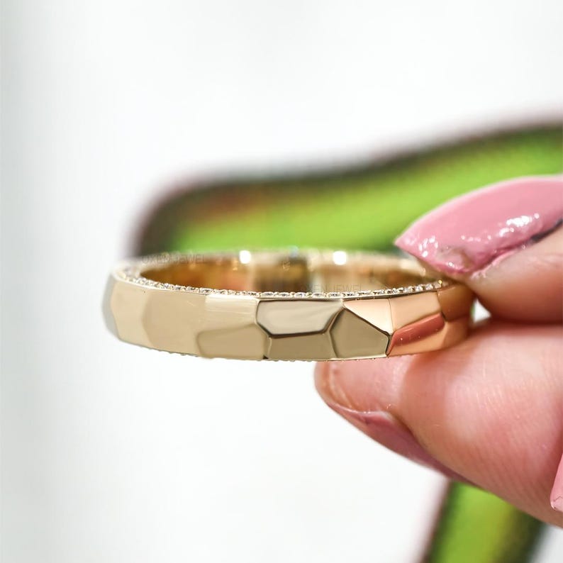 May include: A gold ring with a geometric design. The ring has a faceted surface and a band lined with small, sparkling stones. The ring is held up against a blurred green and white background.