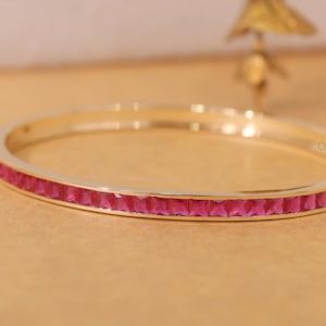 May include: A gold-coloured bangle bracelet featuring a line of square, bright pink gemstones. The bracelet is presented on a light brown surface. The text "Oxen Jewel" is visible on the right side of the image.
