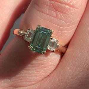 May include: A gold ring featuring a large emerald-cut green gemstone flanked by two smaller baguette-cut diamonds.