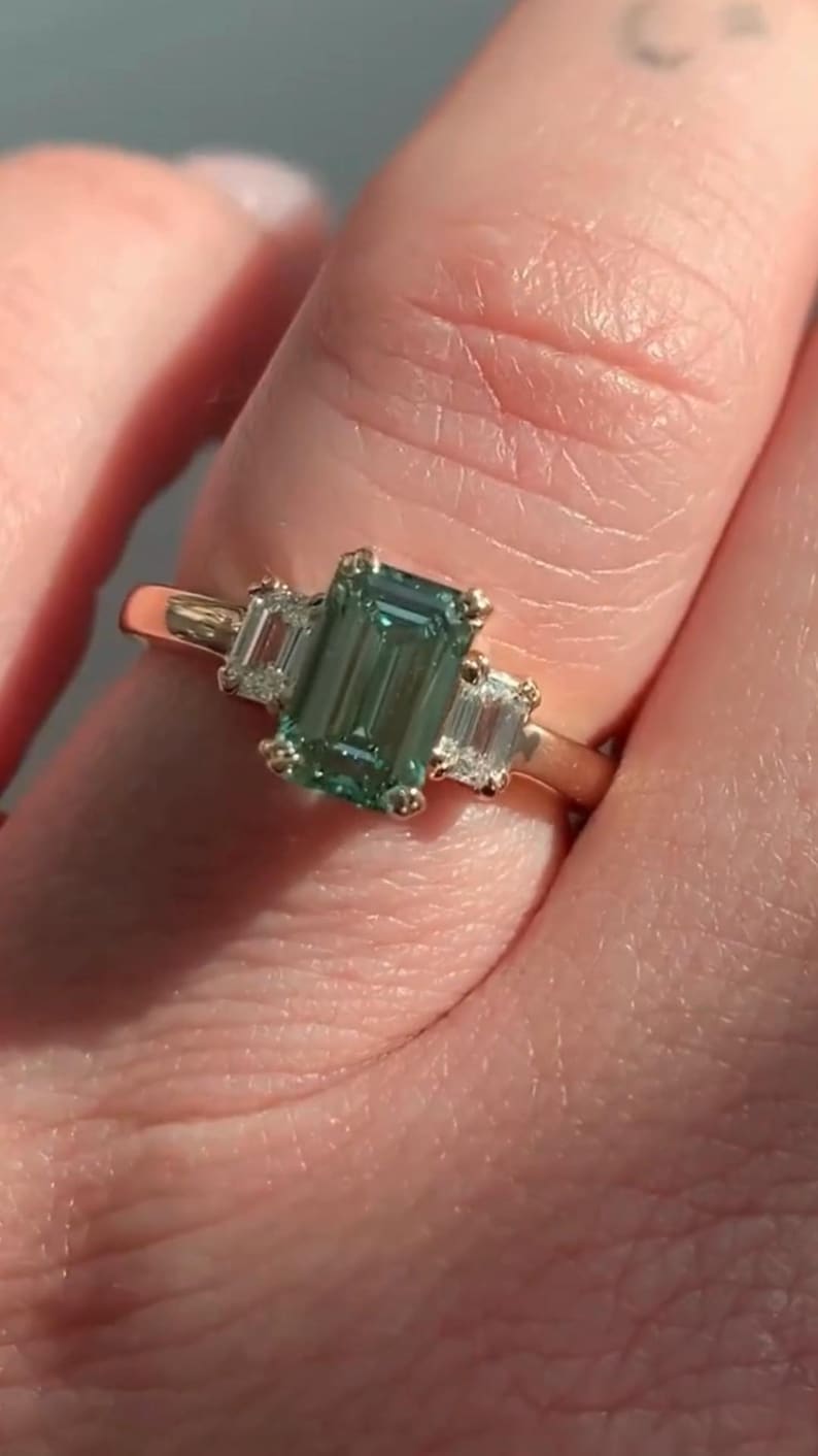 May include: A gold ring featuring a large emerald-cut green gemstone flanked by two smaller baguette-cut diamonds.