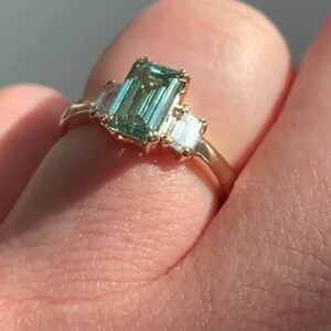 May include: A gold ring featuring a large emerald-cut green gemstone flanked by two smaller baguette-cut diamonds.