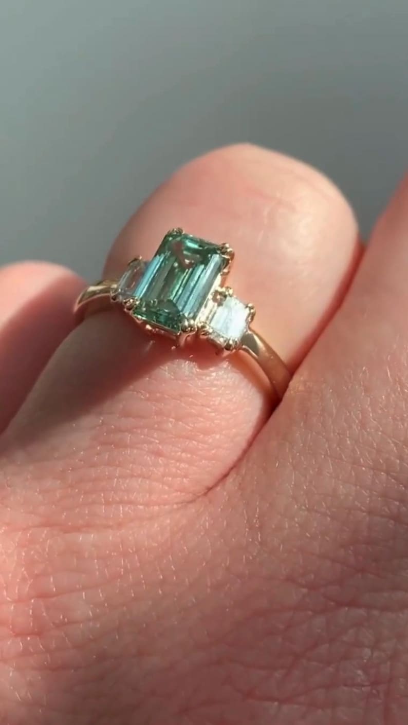 May include: A gold ring featuring a large emerald-cut green gemstone flanked by two smaller baguette-cut diamonds.