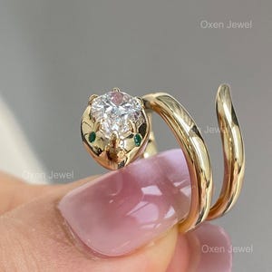 Puede incluir: Anillo de serpiente dorado con una gran gema redonda y transparente en la cabeza. La serpiente tiene dos peque&ntilde;as gemas verdes como ojos. El anillo se enrolla alrededor del dedo en espiral. El texto "Oxen Jewel" es visible.