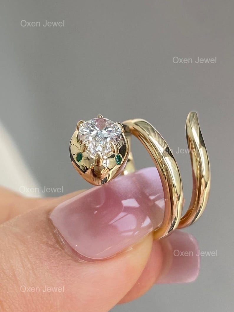 Puede incluir: Anillo de serpiente dorado con una gran gema redonda y transparente en la cabeza. La serpiente tiene dos peque&ntilde;as gemas verdes como ojos. El anillo se enrolla alrededor del dedo en espiral. El texto "Oxen Jewel" es visible.