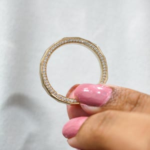 May include: A gold ring with a geometric design, encrusted with small, sparkling diamonds. The ring is held up against a white background, showcasing its intricate details and radiant shine. The ring's band has a faceted design.
