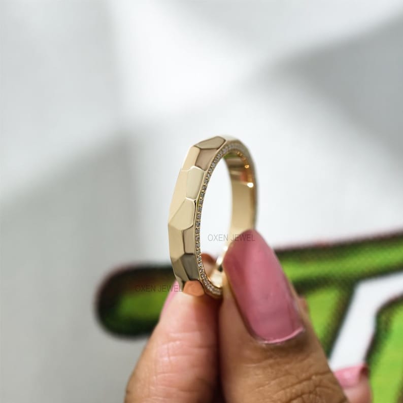 May include: A gold ring with a geometric design. The ring has a series of angled facets and a band of small, sparkling stones. The ring is held up by a person with pink nail polish.