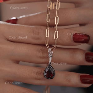 Könnte beinhalten: Eine goldfarbene Halskette mit einem tropfenförmigen Anhänger. Der Anhänger hat einen dunkelroten Edelstein mit schwarzem Rand. Der Text "Oxen Jewel" ist sichtbar. Die Kette wird von einer Person gehalten.