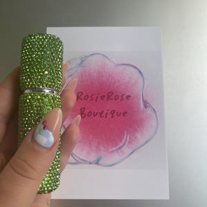 May include: A green, bedazzled perfume atomizer is held in front of a white box with the text "RosieRose Boutique" on it. The atomizer is covered in small, sparkling green gems. The box has a pink watercolour design.