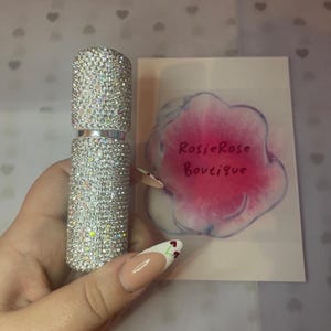 Puede incluir: Un frasco de perfume plateado cubierto de diamantes de imitación. El frasco está siendo sostenido por una mano con arte de uñas de cereza.