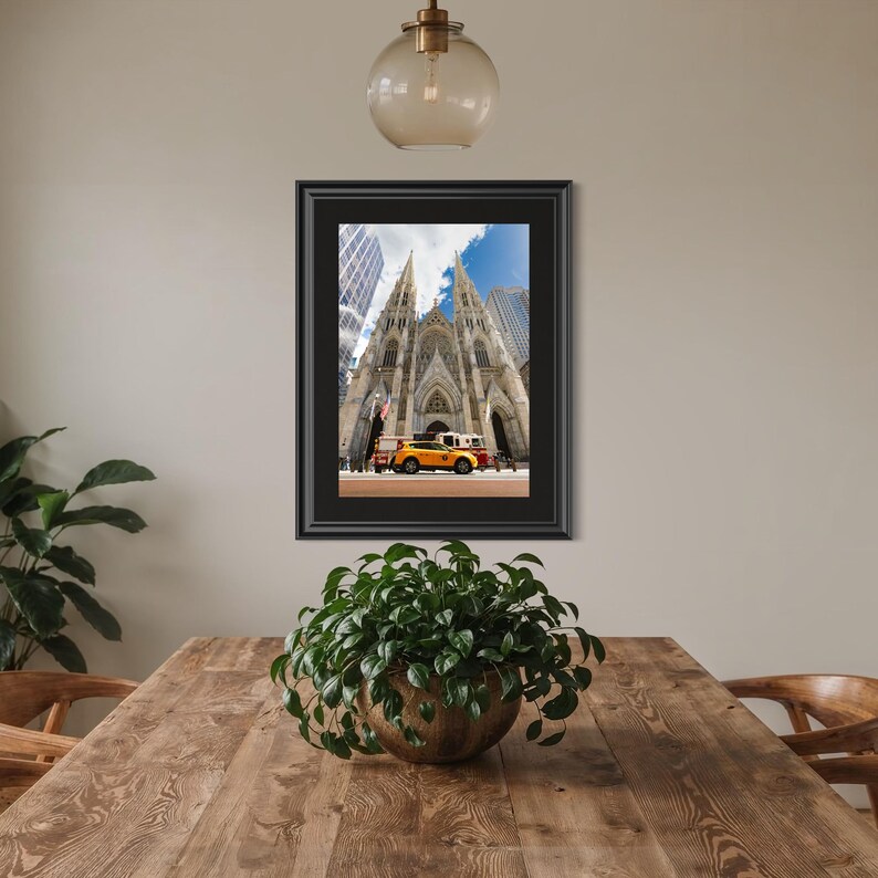 May include: Framed print of St. Patrick's Cathedral in New York City, featuring a yellow taxi cab and other vehicles in front of the cathedral. The image is set against a backdrop of a blue sky and tall buildings.
