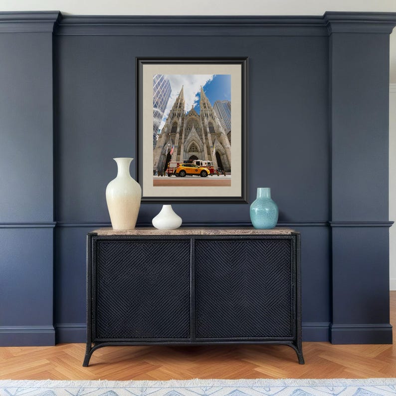 May include: A framed photograph of St. Patrick's Cathedral in New York City, with a yellow taxi cab in front. The photograph is displayed above a black cabinet with a marble top, and three decorative vases.