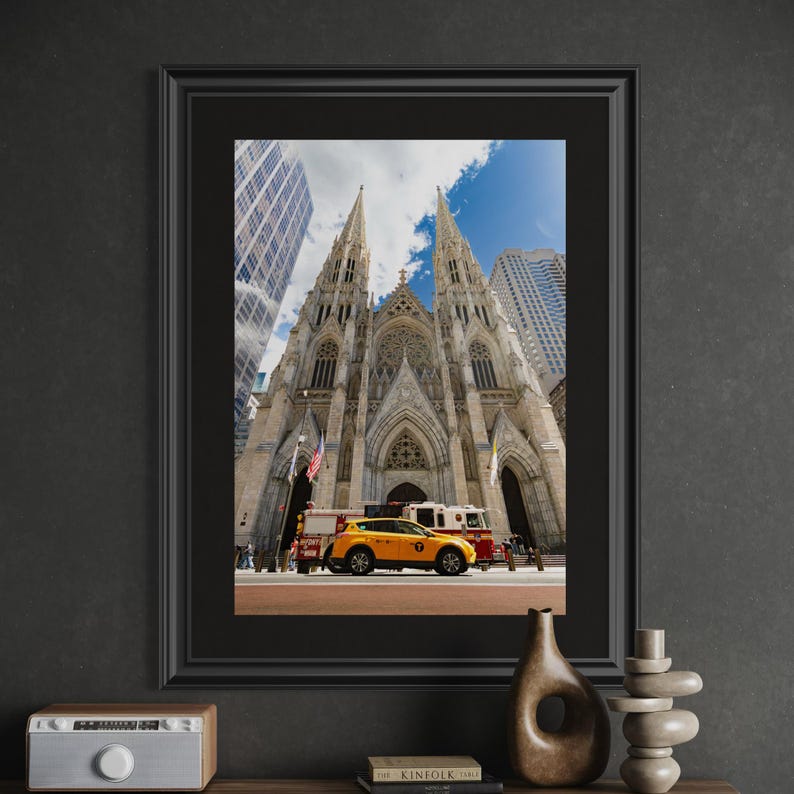 May include: Framed photograph of St. Patrick's Cathedral in New York City, featuring a yellow taxi and fire trucks. The cathedral's spires reach towards a blue sky with clouds, surrounded by skyscrapers.