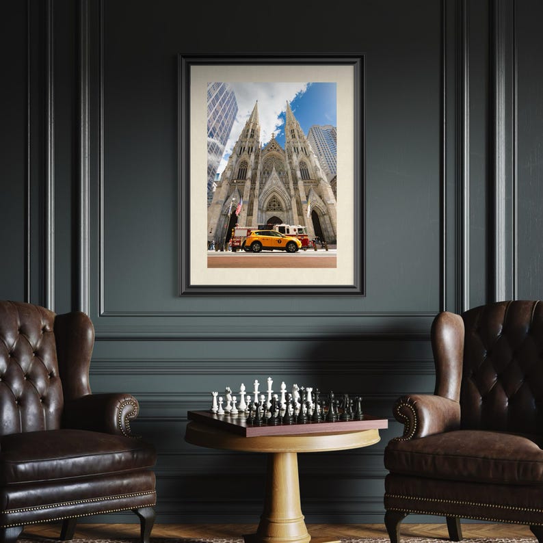 May include: Framed photograph of St. Patrick's Cathedral in New York City, with a yellow taxi cab and other vehicles in front. The image is set in a dark room with two leather armchairs and a chess set on a round table.