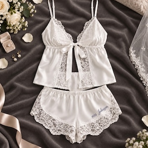 May include: A white satin camisole and shorts set featuring delicate lace trim, adjustable spaghetti straps, and a center front ribbon tie. The shorts are detailed with white lace panels on the sides and the embroidered text mrs Johnson in blue.