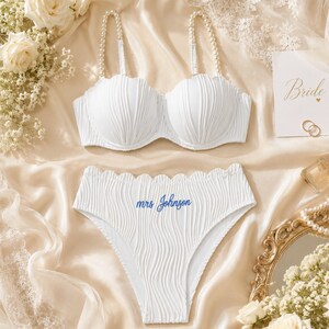 May include: A white two-piece lingerie set featuring a textured, wavy fabric. The bra has pearl-embellished straps and scalloped edges. The high-waisted briefs feature a scalloped waistband and the blue embroidered text "mrs Johnson" on the front.