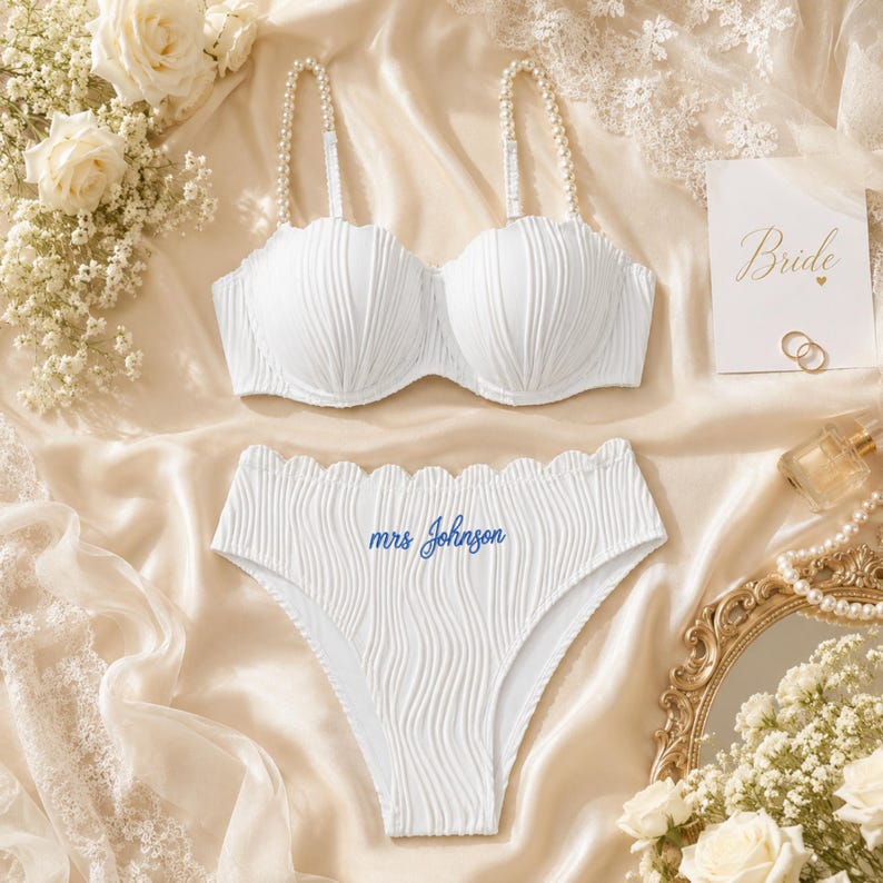 May include: A white two-piece lingerie set featuring a textured, wavy fabric. The bra has pearl-embellished straps and scalloped edges. The high-waisted briefs feature a scalloped waistband and the blue embroidered text "mrs Johnson" on the front.