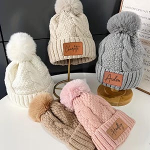 May include: A collection of cable-knit beanies in various colors, including white, beige, gray, brown, and pink. Each hat features a faux fur pom-pom and a leatherette patch with a name. The names "Charlotte", "Aiden", and "Hadley" are visible.
