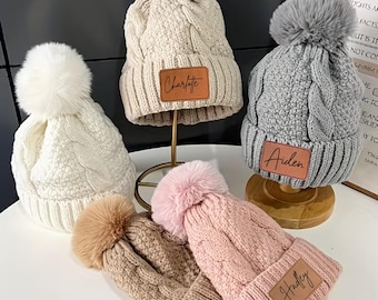 May include: A collection of cable-knit beanies in various colors, including white, beige, gray, brown, and pink. Each hat features a faux fur pom-pom and a leatherette patch with a name. The names "Charlotte", "Aiden", and "Hadley" are visible.