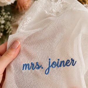 May include: A white sheer mesh fabric featuring delicate lace trim along the top edge. The fabric is embroidered with the text "mrs. joiner" in a blue cursive script font. The material is lightweight, semi-transparent, and has a fine grid texture.