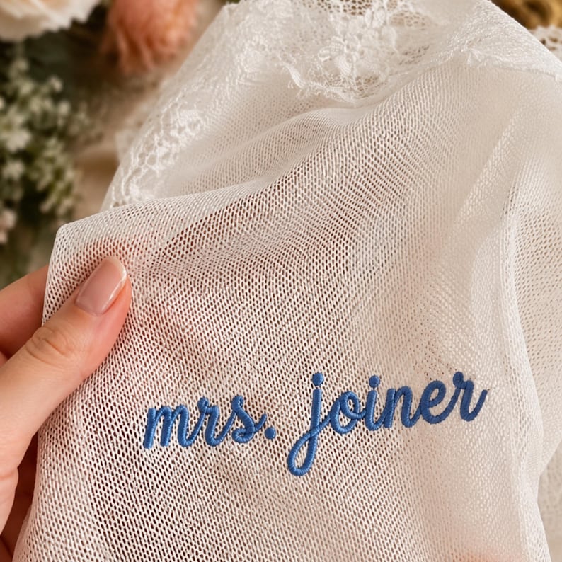 May include: A white sheer mesh fabric featuring delicate lace trim along the top edge. The fabric is embroidered with the text "mrs. joiner" in a blue cursive script font. The material is lightweight, semi-transparent, and has a fine grid texture.