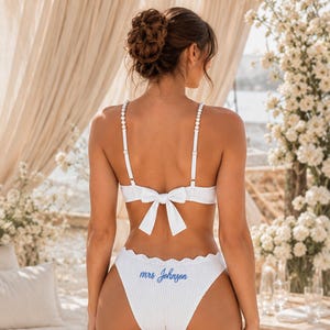 May include: A white ribbed two-piece swimsuit featuring a bikini top with pearl-accented straps and a centre back bow. The high-cut bikini bottom features a scalloped edge and the blue embroidered text "mrs Johnson" on the back.