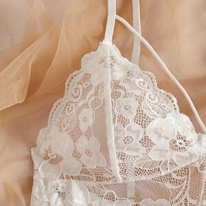 May include: A white lace camisole featuring a delicate floral pattern with scalloped edges along the neckline. The garment includes thin, adjustable white shoulder straps and a sheer, intricate lace overlay that covers the upper bodice section.
