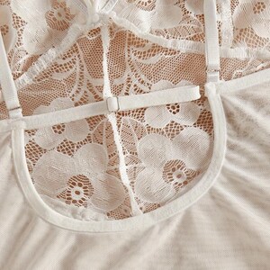May include: A white lace camisole featuring a floral pattern, thin adjustable shoulder straps with silver-toned metal sliders, and a U-shaped cutout detail on the front chest area. The garment is constructed from a sheer, delicate lace fabric.