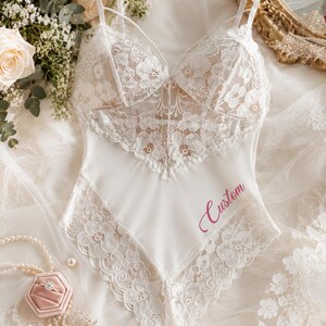 May include: A white bodysuit featuring intricate floral lace detailing on the bodice and leg openings, thin spaghetti straps, and the word Custom embroidered in a script font on the lower right side of the smooth fabric panel.